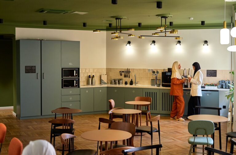 A bright office break room with café tables, green cabinets and two colleagues making coffee at the counter